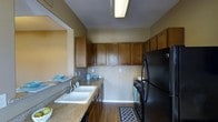 This single-story space centers around a well-appointed kitchen designed for both functionality and comfort. The kitchen features rich, warm-toned wood cabinetry that extends along the upper and lower walls, providing generous storage capacity. Granite-style countertops run the length of the kitchen, offering ample workspace for meal preparation. A double-basin sink is conveniently positioned along the counter, complemented by a standard faucet and additional counter space on either side.

The kitchen is equipped with black appliances, including a full-sized refrigerator, a gas range with oven, and a dishwasher, creating a cohesive and practical cooking environment. Overhead fluorescent lighting ensures the workspace is well-illuminated. Hardwood-style flooring runs throughout, adding warmth to the space.

The kitchen layout follows a galley-style configuration, with counters and appliances lining both sides, creating an efficient workflow. A pass-through or open wall connects the kitchen to an adjacent area, enhancing the overall sense of openness. Neutral warm tones on the walls complement the cabinetry, contributing to an inviting and cohesive atmosphere throughout the space.