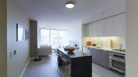 This stylish single-floor space showcases a thoughtfully designed open-concept layout that seamlessly integrates the living, dining, and kitchen areas into one cohesive environment. The kitchen is outfitted with flat-panel cabinetry in a soft grey finish, under-cabinet lighting, a stainless steel sink, and a clean white countertop that reflects the unit's modern aesthetic. A bold dark-topped kitchen island anchors the space, paired with sleek wire bar stools that add an industrial touch. The dining and living areas flow naturally from the kitchen, creating an inviting and functional shared space. A large arc floor lamp serves as a design focal point in the living area, complementing the neutral-toned sofa and understated décor. Floor-to-ceiling windows flood the interior with abundant natural light and offer expansive views of the surrounding cityscape, enhancing the open, airy feel of the space. Light hardwood-style flooring runs throughout, tying the areas together with warmth and continuity. The overall ambiance is clean, contemporary, and sophisticated, ideal for those who appreciate minimalist urban living with high-quality finishes and a bright, welcoming atmosphere.