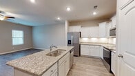 This two-story home is designed with a practical, open-concept layout that connects the kitchen and living areas seamlessly. The kitchen is a standout space, anchored by a central island with a double stainless steel sink and granite countertops featuring warm, neutral tones. White cabinetry with brushed nickel hardware lines the perimeter walls, complemented by under-cabinet lighting and light wood-style flooring that extends throughout the main living area. Stainless steel appliances, including a French door refrigerator with an in-door dispenser, a range, microwave, and dishwasher, complete the kitchen. A pantry door provides additional storage convenience. The adjacent living area is generously proportioned, featuring recessed lighting and a ceiling fan overhead. A staircase leads to the upper level, where carpeted bedrooms offer a comfortable retreat. One bathroom showcases marble-style tile flooring, a single-sink vanity with white cabinetry, and a glass-enclosed walk-in shower. Additional utility space includes an electrical panel area. Neutral wall tones throughout create a cohesive, move-in-ready aesthetic.