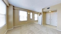 This single-floor space presents a clean, functional layout designed for comfortable everyday living. The main living area is generously sized, finished with wall-to-wall carpeting in a warm neutral tone that complements the beige-painted walls throughout. Three large windows line the exterior wall, allowing ample natural light to fill the room and create a bright, open atmosphere. A wall-mounted light fixture adds a subtle decorative touch to the living space. The textured white ceiling contributes to the overall airy feel of the room. Transitioning toward the back of the space, the kitchen area is visible and features a full-sized refrigerator, with cabinetry overhead providing practical storage solutions. The kitchen is set apart by a tiled floor, offering a clean contrast to the carpeted living area. A white entry door is positioned at the rear, providing access in and out of the unit. A ceiling-mounted HVAC vent ensures climate control throughout the space. The overall design is straightforward and move-in ready, with a cohesive neutral color palette that makes the space feel welcoming and versatile for a variety of furnishing styles.