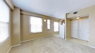 This single-floor space presents a clean, functional layout designed for comfortable everyday living. The main living area is generously sized, finished with wall-to-wall carpeting in a warm neutral tone that complements the beige-painted walls throughout. Three large windows line the exterior wall, allowing ample natural light to fill the room and create a bright, open atmosphere. A wall-mounted light fixture adds a subtle decorative touch to the living space. The textured white ceiling contributes to the overall airy feel of the room. Transitioning toward the back of the space, the kitchen area is visible and features a full-sized refrigerator, with cabinetry overhead providing practical storage solutions. The kitchen is set apart by a tiled floor, offering a clean contrast to the carpeted living area. A white entry door is positioned at the rear, providing access in and out of the unit. A ceiling-mounted HVAC vent ensures climate control throughout the space. The overall design is straightforward and move-in ready, with a cohesive neutral color palette that makes the space feel welcoming and versatile for a variety of furnishing styles.