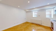 Located at 310 Lonsdale Road in Toronto's Forest Hill neighbourhood, this thoughtfully designed unit offers a clean, modern aesthetic across its single floor. The main living area immediately draws the eye with its striking herringbone-patterned hardwood floors in a warm honey tone, which contrast beautifully against the crisp white walls and smooth white ceiling. Recessed pot lights are positioned throughout, providing consistent, bright illumination. A window with a radiator beneath it allows for natural light and efficient heating. The integrated kitchenette features flat-front white cabinetry, a stainless steel sink, and built-in appliances including a microwave, maintaining a streamlined and functional look that complements the overall minimalist design. A hallway connects the living areas to two well-appointed bathrooms, each finished with large-format neutral grey tiles, a floating white vanity with under-mount sink, a wall-mounted towel radiator, and a large mirror. The consistent use of white tones and quality finishes throughout creates a cohesive, contemporary feel. The space is efficiently laid out, making practical use of every square metre while maintaining an open and uncluttered ambiance.