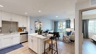 This well-designed single-floor residence offers a cohesive, contemporary aesthetic with a coastal-inspired palette of soft blues, whites, and natural wood tones. The open-concept layout seamlessly connects the kitchen, dining, and living areas, creating a spacious and airy atmosphere filled with natural light.

The kitchen is outfitted with white shaker-style cabinetry, a grey tile backsplash, stainless steel appliances including a refrigerator, range, microwave, and dishwasher, and a white quartz countertop island with bar seating — ideal for casual dining or entertaining.

The living room is bright and inviting, featuring sliding glass doors that open to a private balcony overlooking palm trees, with bold blue drapery adding a stylish coastal accent. Light wood-style flooring extends throughout the main living areas.

Two bedrooms offer comfortable retreats, each with built-in closets and windows framing lush outdoor greenery. The primary bedroom includes a ceiling fan and generous natural light.

The bathroom is finished in crisp white with marble-look tile flooring, a single-sink vanity with a framed mirror, and a tiled tub-shower combination. Throughout the residence, recessed lighting and a clean, neutral color scheme contribute to an overall sense of modern comfort and calm.