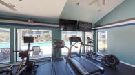 Located at 9001 Jones Rd in Houston, Texas, this fitness center is a well-appointed, single-floor amenity space designed to support a comprehensive workout experience. The interior is characterized by vaulted ceilings with ceiling fans, teal and white accent walls trimmed with bold blue and gray horizontal stripes, and durable rubber flooring throughout — creating an energetic yet functional atmosphere.

The cardio zone is positioned along a wall of large picture windows that frame direct views of the outdoor swimming pool and surrounding greenery, flooding the space with natural light. Treadmills, stationary bikes, and an elliptical trainer are arranged to take full advantage of these views, with wall-mounted flat-screen televisions overhead for entertainment during workouts.

The strength and functional training area is generously equipped with a multi-station cable machine, a weight bench, a dumbbell rack, and a full collection of medicine balls and stability balls on a dedicated rack. A glass-panel partition divides zones within the space while maintaining an open, airy feel.

Glass-paned doors lead directly to the outdoor pool area, enhancing connectivity between indoor and outdoor amenities. The overall design prioritizes both practicality and comfort, making this fitness center a standout feature of the property.