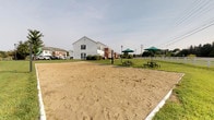 Located at 6340 Saddler Way in Canal Winchester, Ohio, this outdoor amenity space offers a well-appointed community gathering area designed for recreation and relaxation. The centerpiece of the space is a generously sized sand volleyball court, providing a dedicated area for active outdoor play. Adjacent to the court, several picnic tables shaded by green market umbrellas are arranged on a manicured lawn, creating an inviting setting for outdoor dining and socializing. Classic lamp posts punctuate the grounds, adding both functional lighting and a charming aesthetic detail. A white fence runs along the perimeter, lending a clean, defined boundary to the space and contributing to its welcoming character. The surrounding landscape features well-maintained green grass that extends alongside the residential buildings, which are constructed with brick and white siding in a traditional style. The open sky overhead and the spacious layout of the grounds give the area a bright, airy feel. Overall, this community outdoor space balances active recreation with comfortable social areas, making it a functional and attractive amenity for residents of the Canal Winchester community.