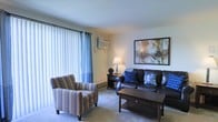 This single-floor space offers a warm and functional layout designed for comfortable everyday living. The living room serves as the central gathering area, featuring wall-to-wall carpeting that adds a soft, welcoming feel underfoot. Large windows dressed with vertical blinds and accent curtains allow natural light to fill the room while providing privacy and light control. A wall-mounted air conditioning unit ensures year-round comfort, complemented by a baseboard heating system along the exterior wall. The living room is furnished with a leather sofa accented by blue throw pillows, a coordinating striped armchair, and a dark wood coffee table, creating a cohesive and inviting seating arrangement. A table lamp on a dark wood nightstand provides warm ambient lighting, while a large framed abstract artwork anchors the wall, adding a decorative focal point to the space. The neutral wall tones throughout provide a versatile backdrop that complements a variety of décor styles. Overall, the space presents a tidy, well-maintained interior with a practical and comfortable ambiance ideal for relaxed living.