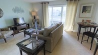 This thoughtfully designed single-floor space offers a comfortable and inviting atmosphere centered around a beautifully arranged living area. The living room is highlighted by a large picture window dressed with flowing cream-toned curtains, allowing generous natural light to fill the room while framing pleasant views of the surrounding neighborhood and greenery beyond.

The space is furnished with a plush, neutral-toned sofa that anchors the seating area, complemented by a classic patterned accent chair that adds visual interest and character. A glass-top coffee table with a dark frame serves as an elegant focal point, accented by a decorative silver candelabra. A matching side table provides additional surface space, while a low-profile media console along the wall accommodates entertainment needs.

Warm, neutral wall tones and soft carpeted flooring contribute to the overall sense of comfort and calm throughout the space. A floor lamp provides ambient lighting, enhancing the cozy atmosphere during evening hours. The layout is open and well-proportioned, allowing for easy movement and a natural flow between furnishings. Overall, this space strikes a balance between functional living and refined aesthetics, making it a welcoming environment suited to both relaxation and everyday living.