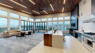 This single-floor amenity space is defined by its striking architectural design, combining warm walnut wood ceiling paneling with sleek linear LED lighting and soaring ceiling heights that create an airy, open atmosphere. Expansive floor-to-ceiling windows wrap around the main living area, flooding the interior with natural light and offering panoramic views of the surrounding landscape and tree line.

The open-plan main area features a large central island with a rich walnut wood countertop and bar-height seating, serving as both a social hub and workspace. Adjacent to this is a fully equipped kitchen outfitted with stainless steel appliances, including a professional-grade range, built-in wine and beverage coolers, a stainless steel range hood, and sleek blue cabinetry topped with white quartz countertops — all accented by a textured white tile backsplash.

The lounge area provides comfortable seating, including a curved velvet sofa paired with a glass coffee table, ideal for casual gatherings. A separate, more intimate media room features a large wall-mounted television, a bold dark textured accent wall, and eclectic modern seating. The overall ambiance blends contemporary design with warm, natural materials, creating an inviting and functional communal environment.
