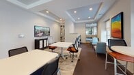 Located at 100 Exchange Street in Malden, Massachusetts, this single-floor amenity space offers a well-appointed, contemporary environment designed for productivity and relaxation. The layout spans a generous open footprint, organized into distinct functional zones that flow seamlessly from one to the next.

The coworking area features modern white tables paired with ergonomic task chairs, recessed lighting, and a tray ceiling detail that adds architectural depth. A wall-mounted flat-screen display serves as a central media focal point, while glass-paneled doors connect to adjacent rooms, maintaining an open and transparent feel throughout.

The kitchen is a standout feature, showcasing a granite-topped island with bar seating, dark espresso cabinetry, stainless steel appliances including a full-size refrigerator and microwave, and large-format tile flooring with a marble-like finish. A secondary kitchenette, also fitted with granite countertops and dark cabinetry, provides additional convenience.

The lounge area is bathed in natural light from oversized windows with roller shades, offering views of the surrounding streetscape. Comfortable seating in blue and orange upholstery creates a welcoming atmosphere. Throughout the space, neutral gray walls, tray ceilings, and consistent flooring materials unify the design, resulting in a polished, professional, and inviting communal environment.