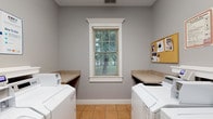 Located at 2422 Banks Edge Way in Reynoldsburg, Ohio, this single-floor space is a well-appointed shared laundry facility designed with both function and comfort in mind. The laundry room is thoughtfully laid out, featuring two full-sized washing machines and two dryers positioned on either side of the room, providing ample capacity for residents. Flanking each appliance are laminate folding countertops that offer convenient surface space for sorting and folding laundry. The room is finished with neutral gray walls and warm beige tile flooring, giving it a clean and welcoming atmosphere. A centrally placed window fitted with white-trimmed molding and horizontal blinds allows natural light to filter into the space while offering a pleasant view of the surrounding greenery. A cork bulletin board mounted on the wall provides a practical community information hub. The crisp white trim and consistent finishes throughout contribute to a tidy, well-maintained environment. Overall, the space is designed to serve as an efficient and comfortable shared amenity, combining practical laundry functionality with thoughtful design details.