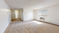 The Nesa Classic is a one-bedroom, one-bathroom unit thoughtfully arranged on a single floor. The living area is generously proportioned for its footprint, featuring wall-to-wall beige carpeting, crisp white walls, and a textured ceiling. A window fitted with horizontal blinds allows natural light to fill the room, while a wall-mounted HVAC unit and baseboard heating ensure year-round climate control. The entry door opens directly into this open living space, providing a straightforward and accessible layout.

The bedroom continues the carpeted flooring and includes a window dressed with vertical blinds that invite natural light, a baseboard heater, and a closet with built-in shelving for storage.

The kitchen features dark wood cabinetry with ample upper and lower storage, light-toned countertops, and a double stainless steel sink. A ceiling-mounted light fixture provides task lighting for the workspace.

The bathroom is finished with white square tile lining the walls of the tub surround, complementing a full-sized soaking tub. A small window above the tub allows for natural ventilation and light. The overall aesthetic throughout the unit is clean, neutral, and functional.