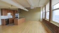 This single-floor loft-style space showcases an open-concept layout that seamlessly integrates living and kitchen areas. The kitchen is outfitted with warm wood cabinetry, a stainless steel refrigerator, a sleek range hood, and a central island with a dark countertop, providing both functionality and style. Exposed cylindrical ductwork runs prominently across the white-beamed ceiling, lending an industrial character to the space. Track lighting is installed along the ceiling, offering flexible illumination throughout. A ceiling fan adds to the practicality of the open layout. Light bamboo or blonde hardwood flooring extends across the entire space, contributing to a clean and airy feel. The walls are painted in a muted olive green, adding warmth and depth to the interior. Large windows along one wall allow natural light to filter in, revealing brick detailing that hints at the building's original structure. The combination of industrial and modern elements creates a distinctive, versatile ambiance suited to a variety of uses.