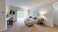 This single-floor residence offers a thoughtfully designed layout that balances comfort and functionality across its interconnected living spaces. The open-plan living and dining area is anchored by plush wall-to-wall carpeting and neutral tones, creating a warm and inviting atmosphere. Large sliding glass doors in the living room flood the space with natural light and provide access to an outdoor balcony, offering pleasant views of surrounding greenery. A wall-mounted television and ample room for seating arrangements make the living area both practical and relaxing. Adjacent to the living space, a dedicated dining area accommodates a full dining table and chairs, seamlessly connecting to the kitchen via a breakfast bar with counter seating. The kitchen features a compact, efficient U-shaped layout with light-toned cabinetry, laminate countertops, stainless steel appliances including a gas range and dishwasher, and a window that brings in additional natural light. Two well-sized bedrooms, both finished with soft carpeting and neutral walls, offer restful retreats with ample space for furnishings. The bathroom is cleanly appointed with a vanity, mirror, and a shower-tub combination. Throughout the residence, a cohesive color palette of beige, cream, and soft teal accents contributes to a calm and comfortable ambiance.