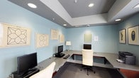 This internet café offers a functional and visually calm environment designed for focused computer use. The single-level space is finished with soft blue walls that create a serene, distraction-free atmosphere. Recessed ceiling lights provide consistent, even illumination throughout, complemented by a classic table lamp that adds a warm accent near one of the workstations.

The room features built-in wraparound desk surfaces along the walls, accommodating multiple computer setups. Workstations are equipped with desktop monitors and keyboards, arranged to maximize available space without feeling cramped. A white ergonomic office chair provides comfortable seating at the central station, while additional seating is visible at adjacent terminals.

The ceiling incorporates a subtle tray design with contrasting gray paneling, adding architectural interest overhead. Security is addressed with visible surveillance cameras mounted at the ceiling. The walls are decorated with several framed geometric artworks, lending a polished, curated quality to the space. Carpeted flooring in a neutral tone completes the interior, contributing to a quiet, professional ambiance suited for browsing, work, or communication.