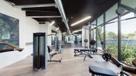 Located on NW 11th Ave in Miami's dynamic urban core, this single-floor fitness center is designed to deliver a premium workout experience. The space features an industrial-modern aesthetic, with exposed ductwork and dark-painted ceilings contrasting against crisp white walls and light hardwood flooring throughout much of the facility.

Floor-to-ceiling glass curtain walls run along the perimeter, flooding the interior with abundant natural light and offering lush, verdant outdoor views of surrounding greenery and palm trees — a distinctly Miami touch. The expansive layout is thoughtfully organized into dedicated zones. The cardio area houses a row of Matrix treadmills, stationary bikes, and elliptical machines positioned directly in front of the large glass facade. A strength and free weights section features adjustable benches, barbell racks, and an extensive dumbbell rack flanked by large wall mirrors. A dedicated functional training zone includes a multi-station cable rig, a heavy punching bag, TRX suspension trainers, and medicine balls.

The facility is accessed through sleek glass pivot doors and includes additional areas visible beyond, suggesting locker room or studio space. Linear LED lighting provides consistent, bright illumination throughout. The overall environment is spacious, well-ventilated, and purpose-built for serious fitness use.