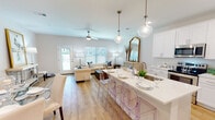 Located at 5054 Via Celeste in Pensacola, Florida, this two-floor home offers a thoughtfully designed layout with a clean, neutral aesthetic throughout. The main level welcomes you through a bright entryway featuring a classic paneled front door with glass inset and a sidelight window that floods the space with natural light. The open-concept living and dining area is anchored by warm wood-look flooring, a ceiling fan, and multiple windows fitted with horizontal blinds that allow for flexible light control. A large kitchen island with a crisp white quartz countertop serves as the centerpiece, accommodating bar seating with modern ghost chairs and pendant lighting above. The kitchen is well-appointed with white shaker cabinetry, stainless steel appliances including a range, microwave, and refrigerator, and generous counter space throughout. The living area flows seamlessly from the kitchen, offering a comfortable and inviting atmosphere. The bathrooms feature white vanities with undermount sinks, light wood-look flooring, and tub-shower combinations. Bedrooms are carpeted for comfort and include ceiling fans, natural light from windows, and generous walk-in closets with wire shelving systems. The overall ambiance is bright, clean, and move-in ready.