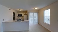 This single-floor space showcases an open-concept layout that seamlessly connects the kitchen and living area. The kitchen is equipped with white cabinetry, a white refrigerator, an electric range, a built-in black microwave, and a double sink set into a white countertop peninsula that separates the kitchen from the living space. The peninsula provides additional workspace and serves as a natural divider between the two areas. The living area is finished with light carpeting and benefits from generous natural light, courtesy of a large sliding glass door fitted with vertical blinds and an additional side window with horizontal blinds. Flush-mount ceiling light fixtures illuminate both the kitchen and living areas. The walls are painted in a neutral beige tone, and the textured ceiling adds a subtle design detail throughout. The overall layout is practical and bright, with an open flow that makes the shared living and kitchen space feel functional and welcoming.