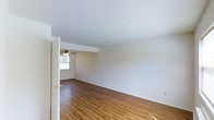 This two-floor space offers a clean, functional layout with a warm and inviting aesthetic. The main living area is spacious and open, showcasing rich hardwood-style flooring that runs throughout and adds a sense of warmth to the neutral, white-painted walls and ceiling. A ceiling fan is centrally positioned, providing both practical airflow and a classic finishing touch to the room. Natural light filters in through a window, brightening the space and creating an airy atmosphere. A baseboard heating unit is visible along the wall, indicating a reliable and efficient heating system integrated into the home's infrastructure. The room's clean lines and simple design make it highly versatile and adaptable to a variety of furnishing styles. Doors with brass hardware provide access to adjacent rooms, maintaining a cohesive and traditional look throughout. The overall ambiance of the space is bright, neutral, and well-maintained, offering a solid foundation for comfortable everyday living across both floors. The straightforward layout and durable finishes reflect a practical yet comfortable design sensibility suited for a range of occupants.