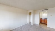 Located along East Route 66 in Flagstaff, Arizona, this single-floor, one-bedroom unit offers a straightforward and functional layout designed for efficient living. The main living area features plush wall-to-wall carpeting that transitions to tile flooring near the entryway, providing a clear distinction between spaces. A front-facing window allows natural light to fill the room, while a wall-mounted light fixture near the entry door ensures adequate illumination. The textured popcorn ceiling and clean white walls give the space a neutral, move-in-ready aesthetic. A built-in open closet with a single hanging rod offers convenient storage within the bedroom area. An arched passageway connects the main living space to an adjoining vanity or wet area, which features warm-toned wood cabinetry, dark countertops, and a wall-mounted light fixture above — adding a touch of character to the otherwise minimal interior. A wall-mounted heating unit near the entry provides climate control. The overall ambiance is clean, simple, and practical, well-suited for individuals seeking a low-maintenance living space in the heart of Flagstaff.