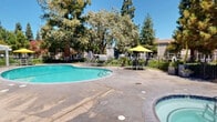 Located at 2424 Hurley Way in Sacramento, California, this outdoor recreational area is a beautifully maintained community amenity space designed for relaxation and leisure. The centerpiece is a generously sized freeform swimming pool with clear, turquoise water, complemented by a separate spa situated conveniently nearby. The pool deck is finished in smooth concrete and is furnished with lounge chairs and seating areas, offering ample space for sunbathing and socializing. A white pergola structure provides a shaded gathering spot, while yellow market umbrellas add vibrant color and additional shade throughout the deck. The space is framed by a wrought iron security fence with a gated entry, ensuring safety and privacy. Surrounding the pool area, lush and mature landscaping creates a serene, park-like atmosphere. Towering redwood and oak trees cast dappled shade across the grounds, with well-tended shrubs, decorative plantings, and manicured greenery lining the pathways. A single-story building with a wood shake roof and dark wood siding borders the space, likely housing support facilities. The combination of vibrant greenery, open blue skies, and thoughtfully arranged amenities makes this outdoor space a welcoming and functional retreat in the heart of Sacramento.