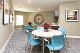This single-floor space in Conyers, Georgia, is designed as a functional and welcoming communal gathering area. The layout is organized around two key zones: a casual seating and dining area and a well-appointed kitchenette, making it well-suited for meetings, gatherings, or collaborative activities.

The seating area features patterned carpet flooring in soft gray tones, lending a comfortable, relaxed atmosphere. Round tables paired with teal-colored chairs create an approachable, social setting. The walls are painted in a warm sage green, accented by framed botanical artwork, decorative wall pieces, and a large statement clock, giving the space a curated yet informal character. A window allows natural light to filter in, brightening the room.

The kitchenette is equipped with rich, dark espresso-toned cabinetry featuring raised-panel detailing and brushed hardware. A light-toned countertop with a stainless steel double-basin sink provides practical workspace, while additional upper and lower cabinetry offers generous storage. A pass-through window connects the kitchenette visually to the seating area, enhancing the open, communicative flow between the two zones. Recessed lighting throughout ensures the space remains well-lit and inviting.