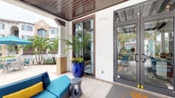 This single-story space presents a well-designed covered entrance that blends indoor comfort with an open-air ambiance. The entryway is defined by a warm, dark wood-paneled ceiling overhead, which adds a refined architectural detail and creates a sense of shelter without enclosing the space entirely. Structural columns frame the area, contributing to its clean, contemporary aesthetic while supporting the covered overhang.

The exterior seating area features a vibrant teal sectional sofa accented with a bold yellow pillow, paired with a modern sculptural side table — offering a welcoming lounge setting for visitors. A striking cobalt blue glazed ceramic planter stands prominently near the entrance, housing a lush tropical palm that adds a lively, resort-like character to the space.

Large steel-framed glass windows and a glass door provide a clear view into the interior, where additional seating and tropical plantings are visible, suggesting a light-filled amenity or lobby area beyond. Light-toned tile flooring runs underfoot, complementing the crisp white exterior walls. The surrounding environment features palm trees and a multi-story building backdrop, reinforcing the tropical, open-air character of this inviting entry space.