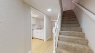 This two-story space is designed with a clean, neutral aesthetic throughout, combining light wood-look flooring on the main level with plush carpeting in the upper-level rooms. The main floor opens into a living area that flows toward a kitchen outfitted with white shaker-style cabinetry, marble-look countertops, a white subway tile backsplash, and stainless steel appliances including a range, built-in microwave, and dishwasher. A pass-through counter connects the kitchen to the living space, enhancing the open feel of the layout. A laundry or utility area with a sink and white cabinetry is also accessible on this level. A staircase featuring a white balustrade and carpeted treads leads to the upper floor, where bedrooms are finished with soft carpet, textured ceilings, ceiling fans, and windows with blinds that allow natural light. The bathroom includes a full tub and shower enclosure with chrome fixtures. A compact bathroom space is also present, tucked beneath the staircase. Throughout, neutral wall tones and consistent trim work create a cohesive, well-maintained environment.