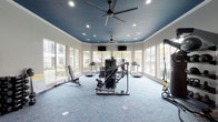 Located at 14723 W Oaks Plaza Dr in Houston, TX, this well-appointed fitness center is designed to support a full range of workout routines in a bright, welcoming environment. The space is defined by its striking deep navy tray ceilings adorned with large ceiling fans and recessed lighting, which contrast elegantly with the light gray walls and blue-toned patterned carpet throughout the main exercise area. An extensive array of large, multi-pane windows lines the perimeter, flooding the space with natural light and providing views of the surrounding landscaped grounds. The main floor is comprehensively equipped with treadmills, a multi-station cable and weight machine, adjustable benches, and TRX suspension training systems. A dedicated section features a heavy punching bag and a weight storage rack for functional training. A secondary area, finished with warm hardwood-style flooring, houses spin bikes, stability balls, resistance bands, and a functional trainer rack, alongside a wall-mounted flat-screen television. The seamless layout accommodates both individual and group workouts, while the open, airy design and abundant natural light create an energizing atmosphere conducive to an active lifestyle.