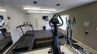 This single-floor fitness center, managed by Woodbury Property Management, offers a practical and well-organized workout space designed to meet essential cardio needs. The room features a rubber flooring mat area designated for equipment use, prominently displaying the Woodbury Property Management branding, while the surrounding floor transitions to a lighter surface, adding visual contrast.

The space is equipped with multiple cardio machines, including two treadmills and two elliptical trainers, providing residents with a variety of workout options. A wall-mounted flat-screen television is positioned for easy viewing during exercise. Overhead fluorescent lighting ensures the room is well-lit, complemented by natural light filtering through windows fitted with blinds.

The walls are finished in a neutral tone, contributing to a clean and functional atmosphere. Ventilation is supported by a ceiling vent, maintaining a comfortable environment during workouts. A small waste bin is conveniently placed near the wall, and a fire extinguisher mount is visible, reflecting attention to safety. The overall ambiance is straightforward and utilitarian, focused on providing a convenient fitness option.