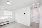 This single-floor unit at 775 Concession Street in Hamilton, Ontario offers a well-maintained, move-in-ready living space defined by a crisp, all-white interior palette and light hardwood-style flooring that runs throughout. The open layout connects the living area to the kitchen via an arched passageway, creating a natural sense of flow between spaces.

The living area is bright and airy, featuring clean white walls, a flush-mount ceiling light, and a baseboard heating unit beneath a window fitted with blinds that allows natural light to filter in. The understated design provides a versatile backdrop for various furnishing arrangements.

The kitchen is compact yet functional, equipped with rich dark wood cabinetry, granite-look countertops, a stainless steel sink, and stainless steel appliances including an electric range and over-the-range microwave. A subway tile backsplash adds a subtle decorative touch.

The bathroom features full white tile surround enclosing a soaker tub with overhead shower, a pedestal-style vanity with a large mirror, and a frosted window that brings in natural light while maintaining privacy.

Panelled interior doors and clean white trim are consistent throughout, contributing to the unit's polished, cohesive appearance.