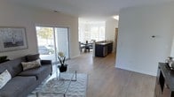 This single-floor space is designed with an open-concept layout that connects the living, dining, and kitchen areas into one cohesive, flowing environment. Light hardwood-style flooring runs throughout, lending a warm, contemporary feel to the entire space. The living area is positioned near large sliding glass doors that allow generous natural light to pour in while providing direct views of the outdoors. A glass-topped coffee table and a neutral-toned sofa complement the clean, modern aesthetic.

The dining area sits centrally within the open plan, illuminated by a stylish chandelier suspended above the dining table. Surrounding windows further brighten the space and enhance the airy ambiance. Adjacent to the dining area, the kitchen features dark cabinetry contrasted against light countertops, and includes stainless steel appliances, including a dishwasher integrated into the island. The kitchen island serves as a natural divider between the cooking and dining spaces while maintaining the open feel.

Throughout the space, crisp white walls, recessed lighting, and consistent flooring create a unified, well-lit environment that feels both functional and inviting.