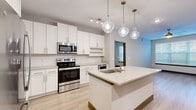 This single-floor residence is designed with a clean, modern aesthetic throughout. The kitchen serves as a focal point, showcasing white shaker cabinetry with brushed nickel hardware, light quartz countertops, and a full suite of stainless steel appliances including an electric range and built-in microwave. A generously sized center island with an undermount sink and gold-tone faucet provides additional workspace and casual seating, while three globe-style pendant lights add a distinctive design element overhead. Track lighting completes the kitchen's well-lit environment.

The bedrooms feature light wood-look flooring, ceiling fans, and large windows fitted with horizontal blinds that allow ample natural light. One bedroom offers views of a surrounding urban streetscape. The primary bathroom includes a dual-sink vanity with quartz countertops, white cabinetry, framed mirrors, and a glass-enclosed walk-in shower. A secondary bathroom mirrors the same refined aesthetic with a single-sink vanity and a shower-tub combination. Both bathrooms connect to walk-in closets outfitted with wire shelving systems. Light wood-look flooring runs consistently throughout the unit, contributing to a cohesive, contemporary feel.