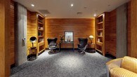 This thoughtfully designed library occupies a single floor and exudes a warm, refined ambiance defined by rich horizontal wood paneling that lines the walls throughout the space. Generous built-in wooden bookshelves are integrated seamlessly into the architecture, displaying an organized collection of books, decorative objects, and curated accessories that lend the space an intellectual character.

The layout is arranged around distinct seating zones, each crafted to encourage different modes of engagement. A central corridor opens into multiple alcoves, where classic leather egg chairs flank a mirrored console table accompanied by a decorative globe and gramophone, creating an intimate and stylish focal point. Elsewhere, a deep leather sofa is paired with an adjustable floor lamp, offering a comfortable setting for extended reading sessions, while additional armchairs and small side tables are positioned throughout.

Soft recessed ceiling lighting casts a warm, even glow across the plush grey carpeted floors, reinforcing the quiet and inviting atmosphere. The interplay between the natural wood tones, leather furnishings, and neutral flooring creates a cohesive and sophisticated aesthetic. An adjacent open area visible through one of the passages hints at a broader facility, suggesting this library serves as a dedicated retreat within a larger complex.
