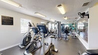 This fitness center is a compact yet comprehensively equipped facility designed to support a full range of workout routines. The space is organized across a single level and is divided into two connected areas, each serving distinct exercise functions.

The main workout area features a lineup of Matrix-brand cardio equipment, including treadmills, elliptical trainers, and stationary bikes, arranged along the perimeter to maximize floor space. A multi-station cable machine anchors the strength training zone, complemented by a dumbbell rack with a full set of free weights and adjustable workout benches. Wall-mounted fans provide additional air circulation throughout.

Full-length mirrors line portions of the walls, creating a sense of expanded space and allowing users to monitor their form. The flooring consists of durable rubber-style material suited for high-traffic fitness use. Natural light filters in through multiple windows fitted with blinds, while overhead fluorescent fixtures ensure consistent illumination throughout the day.

The walls are painted in a soft teal accent color, adding a fresh, energizing atmosphere to the space. A wall-mounted television is present for entertainment during workouts. A glazed interior window and door connect the fitness area to an adjacent lounge or common area, enhancing the open feel of the overall facility. The textured ceiling completes the functional, well-maintained character of this fitness center.