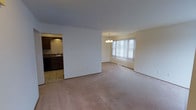 This single-floor residence offers a practical and comfortable layout designed for everyday living. The main living area features wall-to-wall carpeting in a neutral tone, providing a warm and inviting foundation throughout the space. An open flow connects the living room to the dining area, which is accented by an elegant chandelier hanging above and a large window dressed with vertical blinds that allows abundant natural light to fill the room. The bright, airy atmosphere is enhanced by clean white walls and standard-height ceilings throughout. Adjacent to the main living space, the kitchen is finished with tile flooring, dark cabinetry, and a sink positioned beneath recessed lighting, offering a functional workspace that is visually distinct from the carpeted living areas. The seamless transition between the kitchen and living spaces makes the layout feel cohesive and well-connected. Overall, the space presents a clean, move-in-ready interior with a straightforward design that prioritizes openness and functionality.