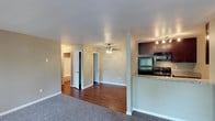 Located at 9505 Gold Coast in San Diego, this upstairs unit offers a thoughtfully designed single-floor layout that balances comfort and functionality. The open-concept living area features plush carpeting that transitions seamlessly into rich hardwood-style flooring, creating a warm and inviting ambiance throughout. A ceiling fan centered in the main living space provides both comfort and a subtle design accent.

The kitchen is semi-open, separated from the living area by a half-wall breakfast bar topped with granite-style countertops. Dark espresso cabinetry lines the kitchen walls, complemented by black appliances including a refrigerator and range, as well as recessed track lighting overhead that keeps the space well-lit and functional.

Two doorways visible from the main living area lead to additional rooms, one of which reveals a spacious walk-in or reach-in closet, suggesting well-appointed bedroom accommodations. The neutral, light-toned walls throughout the unit provide a versatile backdrop suitable for a variety of interior styles.

Overall, the space presents a clean, move-in-ready environment with practical finishes and a cohesive design that makes efficient use of its layout — ideal for those seeking a comfortable residential unit in San Diego.