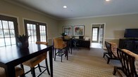 This single-floor space offers a thoughtfully designed interior that blends comfort with functionality. The main area is anchored by a patterned, neutral-toned carpet that adds texture and warmth throughout. A casual seating arrangement features a dark wood high-top table paired with tan upholstered bar-height chairs, creating an inviting spot for casual meetings or collaborative work. Adjacent to this is a dedicated workstation area complete with a dark wood desk, a built-in computer setup, and an ergonomic office chair — ideal for productivity tasks. The walls are finished in a soft, muted green tone accented by crisp white crown molding and chair rail details, lending the space a polished, professional character. Two framed artworks hang above the workstation, adding a decorative touch. Natural light filters generously into the room through multiple windows with classic grid-pane detailing and dark-framed glass doors, connecting the interior to the outdoors. A textured white ceiling with recessed lighting completes the refined aesthetic. The overall ambiance is clean, welcoming, and well-suited for both work and social interaction.