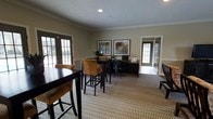 This single-floor space offers a thoughtfully designed interior that blends comfort with functionality. The main area is anchored by a patterned, neutral-toned carpet that adds texture and warmth throughout. A casual seating arrangement features a dark wood high-top table paired with tan upholstered bar-height chairs, creating an inviting spot for casual meetings or collaborative work. Adjacent to this is a dedicated workstation area complete with a dark wood desk, a built-in computer setup, and an ergonomic office chair — ideal for productivity tasks. The walls are finished in a soft, muted green tone accented by crisp white crown molding and chair rail details, lending the space a polished, professional character. Two framed artworks hang above the workstation, adding a decorative touch. Natural light filters generously into the room through multiple windows with classic grid-pane detailing and dark-framed glass doors, connecting the interior to the outdoors. A textured white ceiling with recessed lighting completes the refined aesthetic. The overall ambiance is clean, welcoming, and well-suited for both work and social interaction.
