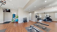 This fitness center offers a spacious, single-floor layout designed to accommodate a comprehensive range of workout activities. The interior features warm hardwood-style flooring throughout, neutral-toned walls, and recessed lighting that creates a clean, energizing atmosphere. Ceiling fans are installed to ensure comfortable airflow during workouts.

The facility is divided into functional zones. The cardio area is well-stocked with treadmills, elliptical trainers, and a rowing machine, all positioned beneath a wall-mounted flat-screen television for entertainment. A dedicated strength training section houses a multi-station cable machine, weight bench, and a power tower for bodyweight exercises. Free weights, including a full rack of dumbbells and stability balls, occupy a separate corner, supported by large wall mirrors that expand the sense of space and allow users to monitor their form.

Natural light flows in through multiple windows and glass-panel entry doors, keeping the environment bright and inviting. The space benefits from multiple entry points, providing easy accessibility. Protective rubber floor mats are strategically placed beneath equipment. Additional amenities include a body weight scale and informational signage. Overall, this fitness center is thoughtfully organized to support both cardio and strength training needs in a comfortable, well-lit setting.