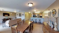 This single-floor space offers an open-plan layout that seamlessly connects a dining area, lounge, and kitchen, creating a versatile environment well-suited for group gatherings. The dining area features a large wooden table surrounded by matching wooden chairs, providing ample seating for multiple guests. Flush-mount ceiling light fixtures illuminate the space evenly, while a ceiling fan in the lounge area adds to the comfort of the room.

The lounge area is positioned adjacent to the dining space and includes a sofa, contributing to a relaxed, communal atmosphere. A wooden dresser or storage cabinet along one wall adds functional storage to the space.

The kitchen is situated at the rear and is equipped with light wood cabinetry, a black refrigerator, a microwave, and countertop appliances. A raised bar counter with industrial-style adjustable bar stools separates the kitchen from the dining and lounge areas, offering additional casual seating. Pendant lights hang above the bar counter, adding a subtle design detail.

The neutral-toned walls and patterned carpet flooring throughout contribute to a warm, welcoming ambiance, making this space practical and comfortable for everyday communal use.