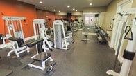 This fitness center offers a comprehensive workout environment designed to accommodate a variety of exercise routines. The space is outfitted with a solid selection of professional-grade equipment, including Quantum and Duo-Tek strength machines, cardio equipment such as ellipticals and treadmills, and a dedicated free weights area featuring a dumbbell rack with accompanying benches. The durable rubber flooring provides both safety and comfort underfoot, while the full-wall mirror installation along one side of the room enhances the sense of openness and allows users to monitor their form during workouts. Recessed lighting is distributed evenly across the ceiling, ensuring the space is well-lit throughout. A bold accent wall in deep orange adds an energizing visual element to the interior, contrasting with the neutral tones of the surrounding walls. A window along the far wall allows for a measure of natural light to enter the space. The layout is thoughtfully organized, providing ample floor space between equipment to allow for safe and comfortable movement. Overall, the fitness center presents a functional and motivating environment suited for a wide range of fitness goals.