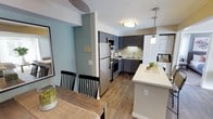 This single-floor residence offers a thoughtfully designed layout that seamlessly integrates the kitchen and dining areas into a cohesive, open-concept living space. The kitchen is a standout feature, outfitted with gray shaker-style cabinetry, stainless steel appliances including a refrigerator, range, and built-in microwave, as well as a deep basin sink beneath under-cabinet lighting. A peninsula with a clean white countertop extends into the space, providing additional prep area and casual seating opportunities, and is accented by a stylish pendant light overhead. The warm hardwood-style flooring runs throughout, adding continuity and a welcoming feel to the interior. Soft, neutral wall tones in sage green and warm beige create a calm and inviting ambiance. The dining area accommodates a full dining table and chairs, making it well-suited for everyday meals or entertaining. A stacked washer and dryer unit is conveniently integrated into the space, adding a practical touch. Flush-mount ceiling lighting ensures the entire area is well-lit, contributing to a bright and functional environment throughout this comfortable, move-in-ready home.