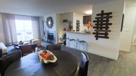 This well-appointed single-floor residence showcases an open-concept design that seamlessly connects the dining, living, and kitchen areas into one cohesive and inviting space. The dining area anchors the layout with a bold, dark round table paired with upholstered chairs, topped with tasteful decorative accents. Rich dark hardwood flooring runs throughout, lending warmth and continuity across the entire space.

The living room is centered around a tile-surround fireplace, creating a comfortable and welcoming focal point. A decorative starburst mirror adorns the wall nearby, adding a touch of contemporary flair, while natural light filters in through a window, brightening the room.

Transitioning toward the kitchen, a raised bar-height counter with sleek metal stools provides casual seating and defines the space without closing it off. Dark cabinetry is visible in the kitchen area above, complemented by open shelving that displays decorative vases and floral arrangements. Wall-mounted art pieces, including a distinctive geometric wood panel, contribute to the space's modern aesthetic throughout. The overall ambiance is stylish and cohesive, blending functional design with contemporary décor elements.