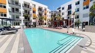 The outdoor amenity area at Eleven10 Apartments at 1110 Town & Country presents a well-appointed, resort-inspired environment designed for relaxation and leisure. The centerpiece is a generously sized swimming pool with crystal-clear turquoise water, framed by a spacious concrete sun deck lined with modern chaise lounges and shaded seating areas. Tall palm trees and a pergola structure with retractable umbrellas add a tropical, comfortable ambiance to the poolside setting.

Adjacent to the main pool, a dedicated spa area features built-in water jets and a sleek, minimalist design, bordered by polished river rock landscaping that lends a clean, contemporary aesthetic. Glass panel barriers provide subtle separation between zones while preserving sightlines. A striking outdoor fireplace with a modern, recessed design and river rock detailing serves as a focal point in the lounge area, complemented by warm wood-slatted privacy screens and drought-tolerant plantings.

The surrounding multi-story residential building features bold color-block accents, expansive balconies, and large windows, creating a dynamic urban backdrop. The overall atmosphere combines modern architectural design with resort-quality outdoor living, all set beneath abundant natural sunlight.
