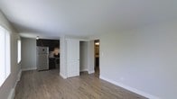 This single-floor space offers a clean, open-concept layout that seamlessly connects the living area to the kitchen. The flooring throughout is a warm-toned, hardwood-style laminate that adds a modern yet inviting feel to the space. The kitchen is equipped with dark espresso-toned cabinetry, stainless steel appliances including a refrigerator and range, and a tile backsplash that provides a polished, contemporary finish. A built-in closet with a classic paneled white door and a metal hanging rod is conveniently positioned between the main living area and the hallway, offering practical storage. Adjacent to the closet, a hallway leads to a bathroom, visible in the background with its own lighting fixture. The walls are finished in a neutral, light tone that enhances the brightness of the space and complements the darker kitchen elements. Clean white baseboards frame the perimeter, contributing to a crisp, well-maintained aesthetic. The overall layout is functional and straightforward, making efficient use of the available space while maintaining a cohesive, modern design throughout.