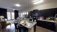 Located at 5225 Las Colinas Blvd in Irving, TX, this single-floor residence offers a thoughtfully designed open-concept layout that seamlessly connects the kitchen, dining, and living areas. The kitchen is a standout feature, outfitted with dark espresso cabinetry, granite countertops, a mosaic tile backsplash, and a full suite of stainless steel appliances including a range, microwave, and refrigerator. A large central island with seating serves as both a functional workspace and a casual dining spot, illuminated by a modern pendant light fixture. Wood-style flooring runs throughout the main living areas, adding warmth to the contemporary aesthetic. The living and dining space is bright and open, benefiting from large windows that invite natural light and provide outdoor views. The bedroom is a comfortable retreat, featuring plush carpeting, a ceiling fan, and oversized windows dressed with blinds and dramatic floor-length drapes. An en-suite bathroom includes a glass-enclosed shower and direct access to a generously sized walk-in closet equipped with built-in shelving and organizational systems. Throughout the space, elevated details such as tasteful wall décor and designer lighting fixtures contribute to a polished, modern ambiance.