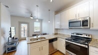 Located at 1702 N Ruddell St in Denton, TX, this well-designed one-bedroom, one-bathroom unit offers a smart and functional layout across a single floor. The open-concept kitchen anchors the living space and is outfitted with white shaker cabinetry, granite countertops, a double-basin sink, and a full suite of stainless steel appliances including a refrigerator, electric range, microwave, and dishwasher. Pendant lighting over the kitchen peninsula adds a modern touch, while hardwood-style flooring flows throughout the main living areas, creating a cohesive and clean aesthetic. The living area benefits from natural light through multiple windows and is equipped with a ceiling fan for year-round comfort. The bedroom features plush carpet, elevated ceilings, and large windows that invite abundant natural light and outdoor views. A dedicated closet provides built-in shelving for additional storage. The bathroom offers a granite-topped vanity with white cabinetry, a large mirror, and a tiled shower-tub combination. A dedicated laundry area adds everyday convenience. Neutral tones and clean finishes throughout give the space a bright, move-in-ready feel well-suited for comfortable single-occupancy living in Denton.