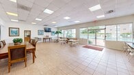 Located at 14759 NE 18th Ave in North Miami, Florida, this single-floor community room offers a generously proportioned, open-plan space designed to accommodate a variety of gatherings and activities. The interior is finished with neutral-toned ceramic tile flooring throughout, contributing to a clean, low-maintenance aesthetic that suits high-traffic use.

The drop ceiling incorporates a grid of recessed fluorescent panels, providing consistent, well-distributed artificial lighting across the entire room. Large windows fitted with horizontal blinds line multiple walls, allowing abundant natural light to filter in while maintaining privacy and light control. The windows also offer views of the surrounding greenery and outdoor areas.

A dedicated computer workstation area runs along one wall, furnished with multiple desktop computers and seating — a practical amenity for residents or members. Additional seating arrangements include folding tables and chairs for flexible use, as well as a formal dining table with upholstered chairs. Framed artwork and a wall-mounted flat-screen television add a welcoming touch to the otherwise functional space.

Double glass doors open directly to an outdoor pool area, seamlessly connecting indoor and outdoor environments and enhancing the room's overall sense of openness.