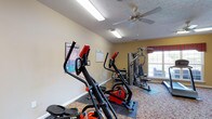 The fitness center at Blankenbaker Crossings is a well-equipped, single-floor facility designed to support a range of workout routines. The room features warm yellow walls, a textured ceiling, and patterned carpeting throughout, creating a welcoming environment. Ceiling fans and fluorescent lighting ensure the space remains well-lit and comfortable during use. A large wall-mounted mirror spans one side of the room, visually expanding the space and allowing users to monitor their form. Natural light filters in through multiple windows dressed with red valances, offering views of the surrounding exterior. The cardio section includes two elliptical cross-trainers and a treadmill, each positioned on protective floor mats. A multi-function cable weight machine and a weight bench provide strength training options. Additional equipment includes a stability ball and a rowing attachment. Workout reference charts and motivational posters are displayed on the walls, along with a posted set of fitness center rules. Near the entrance, a small storage shelf holds sanitizing supplies, reinforcing a clean and organized atmosphere. A wall-mounted television is positioned for convenient viewing during workouts. The layout is efficient and functional, making the most of the available space for a comprehensive fitness experience.