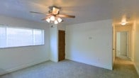 This single-story space presents a clean, straightforward layout highlighted by neutral white walls and wall-to-wall carpeting in a light, neutral tone. The room is generously proportioned, offering an open and airy feel with ample space for a variety of furniture arrangements. A five-blade ceiling fan with an integrated light fixture serves as a functional focal point overhead, providing both ventilation and ambient lighting. Natural light filters into the room through a window fitted with horizontal blinds, brightening the interior while allowing for privacy control. A distinctive louvered wooden door adds a classic architectural detail, providing character and visual warmth against the otherwise minimal backdrop. Clean white baseboards frame the perimeter of the room, contributing to the tidy, well-maintained aesthetic. A wall-mounted control panel is also visible, adding a practical element to the space. The overall ambiance is calm and neutral, making it a versatile environment well-suited for adaptation to various uses or personal design preferences.